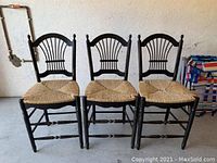 Three black wooden counter stools with woven seats, arranged side by side against a wall with visible scratches on legs and seat support.