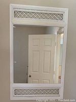 White framed wall mirror with lattice decorative panels at top and bottom, reflecting a partially open door.