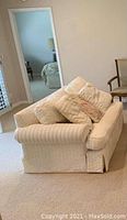 Small cream-colored sofa with quilted fabric upholstery, rolled arms, and skirted base, including three cushions consisting of two large matching quilted cushions and one smaller floral-pattern cushion.