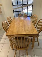 Rectangular wooden kitchen table with six matching spindle-back chairs placed around it in a tiled kitchen space with window blinds.