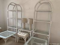 Photo showing two tall white wicker bookcases with arched tops, a cushioned bench/stool, two square glass-top side tables, and a small rectangular glass-top table arranged near a wall.