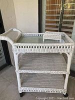 Photo of white wicker serving cart with three shelves and glass tray on top, placed outdoors on a tiled floor near window and wall.