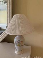 Photo shows the lamp with a white ceramic base decorated with floral designs and a beige pleated fabric shade, placed near a mirror reflecting part of the room.