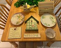 Photo of assorted kitchen decor items arranged on a wooden table including decorative plates, a watering can, a birdhouse, a welcome sign, a wooden organizer, and a pot.