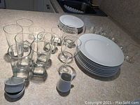 Photo showing a set of white ceramic dinner plates, smaller plates stacked behind, and an assortment of clear glassware including tall glasses, smaller glasses, and two stemmed wine glasses, plus a white ceramic sauce pitcher and a small ramekin.