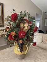 Photo of a faux flower arrangement approximately 3 feet tall with red, pink, and peach artificial flowers, green leaves, and faux grapes, in a yellow and natural colored ceramic vase placed in a living room setting.