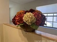 Photo of a faux flower arrangement with burgundy, orange, and cream flowers in a brown vase on a beige wall partition.