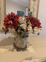 Photo showing artificial flower arrangement with red, cream, and yellow flowers, green leaves, set in decorative ceramic vase with metal stand, placed on a table in front of mirrors.