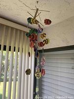 Ceiling mobile hanging from a textured ceiling in front of a window with vertical blinds. The mobile includes multiple colorful fish figures hanging from wooden sticks.