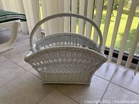 White wicker magazine holder with handle sitting near a window, showing semi-circular design and woven construction detail.