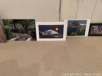 Seven pictures lined up on floor against a wall showing various landscapes and cityscape scenes