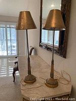 Photo showing two matching tall metal table lamps with gold-toned conical shades and decorative bases placed on a marble tabletop in front of a mirror.