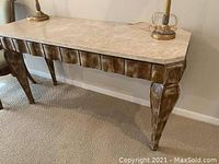 Console table with light marble top and dark carved wooden base photographed against a beige wall with two brass lamps on it.