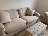 Beige pull out sofa with two seat cushions, two back cushions, two throw pillows, and matching ottoman nearby. Fabric upholstery shows visible wear and light stains.