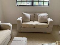 Photo of small beige fabric sofa with two seat cushions, two back cushions, and two throw pillows under a window.
