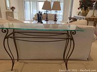 Glass console table shown from side angle against a white sofa, highlighting clear glass top and dark curved metal base.
