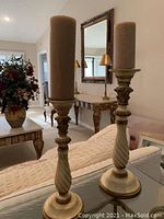 Pair of tall, twisted design candle holders each with a beige pillar candle on a couch.