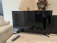 Samsung flat screen television on a beige table with remote controls visible on top, lamp and floral arrangement in background.