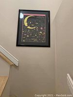 Framed wall art showing a stylized crescent moon with a face above a colorful nighttime cityscape, hung on a beige wall near stairs.