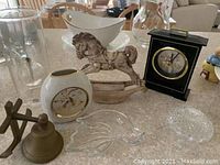 Image shows a collection of decorative items including ceramic rocking horse, oval vase, black desk clock, brass bell, clear glass serving dish and cut glass bowl, and tall glass vase on a countertop.