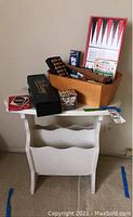 White wooden magazine rack with curved compartments, worn paint visible on legs and edges. Various boxed games in wooden box placed on top including backgammon and card decks.