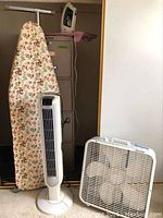 Photo showing floral ironing board standing next to a white tower fan and a white box fan on the floor in front of it, with a metal filing cabinet in the background.