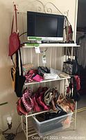 Overview of large lot of handbags, purses, fanny packs and totes arranged on a metal shelving unit with a television on top. Lot contains mostly handbags in various styles, colors and sizes, many vintage with visible bead and sequin details.