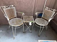 Two white wrought iron chairs flanking a square wrought iron mesh table, placed on concrete floor by wall