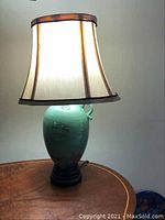 Lamp angled to show the full lamp including the beige fabric lampshade with black trim and green ceramic base with wood platform.