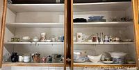 Full view of two kitchen cabinets showing shelves with assorted dishes, plates, cups, and small serving dishes on three shelves.