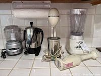 Front view of countertop appliances including a small food processor, electric kettle, immersion blender with stand, on tiled kitchen counter.