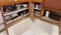 View of open lower kitchen cabinets showing various metal pots, pans, baking dishes, trays, and plastic containers.