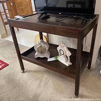 Wooden rolling serving cart holding two decorative clocks on lower shelf. Top shelf has raised edges and a TV and remote on it. Carpeted room.