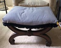 View of foot stool showing dark wood curved base and cushion seat with blue and white checkered fabric with black fringe trim.