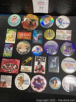 Photo shows 24 assorted colorful button pins arranged on black surface, featuring Disney characters, slogans, and movies.
