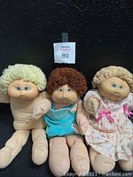 Full view of all three Cabbage Patch Dolls—one nude, one dressed in turquoise onesie, the other in pink floral dress.