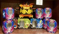 Front view of eight Pokémon-themed metal storage tins arranged in two rows with the Emboar plush in the center back. Tins show colorful character art and windows.