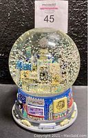 Front view of the musical snow globe showing detailed 3D buildings and snow with glitter inside the globe and colorful billboards on the base.