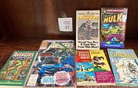 Photo of five paperback books and comic books arranged on a wooden shelf, including Avengers, Hulk, Tarzan, National Lampoon, and Cracked magazine