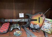 Wide view of video gaming lot items on wooden shelf including Guitar Hero controller, Xbox controller, PlayStation 2 controllers, GTA games, and carrying case.