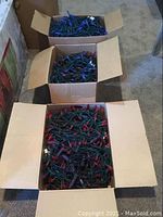 Three open cardboard boxes with assorted tangled red and purple LED Christmas lights on green wiring placed on carpet flooring.