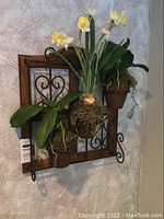 Front angled photo of the iron and wood wall hanging planter with three potted plants, showing decorative iron scrolls and wooden framing, with yellow and white artificial flowers.