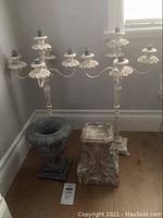 Photo of two white metal iron candelabras, a decorative urn-shaped vase, and a square-based candle stand grouped on floor near wall.