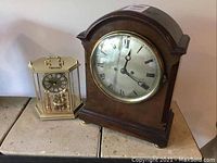 Two clocks side by side: the larger wooden mahogany mantle clock and the smaller decorative plastic clock with gold-tone finish and black dial.