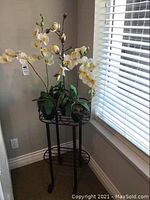 Full view of black iron plant stand with pots and artificial orchid and lily plants beside window