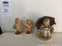 Three Hummel figurines displayed together on a white surface: two small child figurines and one larger girl with umbrella figurine.