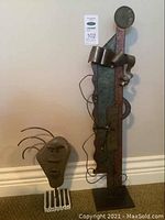 Both metal art pieces side by side showing tall abstract metal art with stone face and clock and smaller face sculpture with wire hair.