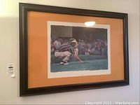 Framed signed limited edition art print of golfer Fred Couples crouching on a green with autograph and number 78/150 in an orange matte and dark wooden frame.