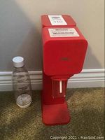 Front view of red SodaStream device with clear plastic bottle next to it on carpet.