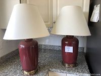 Two matching burgundy ceramic base table lamps with white bell-shaped shades on granite countertop.